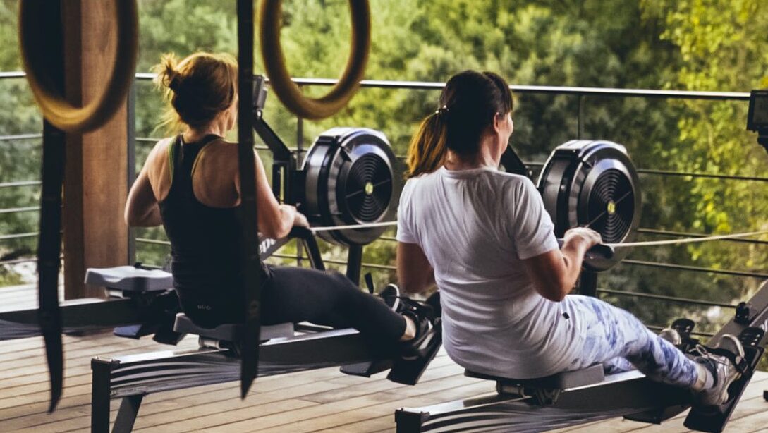 Two members on rowing machines beneath gymnastic rings on a covered outdoor deck