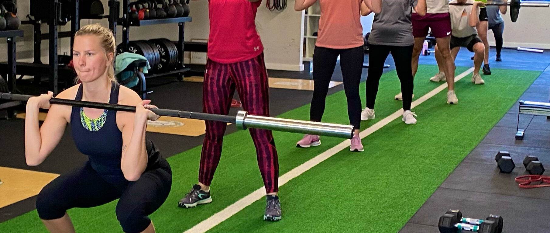 A group class in the studio, one member front squatting a loaded barbell while others train behind her on the green floor